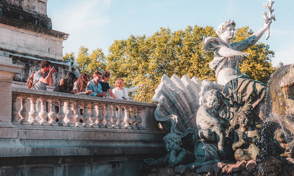Fontäne mit Wasserspielen und antiken Statuen in einem Park in Bordeaux, mit Besuchern auf einer Steinbalustrade im Hintergrund und grünen Bäumen