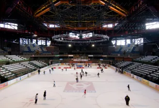 Innenansicht der Eislaufhalle Mériadeck in Bordeaux mit mehreren Schlittschuhläufern auf dem Eis, leeren Rängen und Stahlbachendachkonstruktion mit orange- und gelber Beleuchtung.