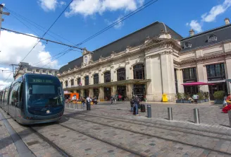 Eine moderne blaue Straßenbahn fährt in den Bahnhof Saint-Jean in Bordeaux ein, vor dem beeindruckenden historischen Gebäude in Beigefarben mit verzierter Fassade und Schieferdach unter blauem Himmel.
