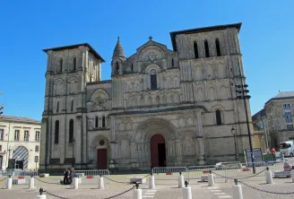 Die Abteikirche Sainte-Croix in Bordeaux, eine romanische Kirche mit zwei quadratischen Türmen, ornamentierter Steinfassade und Hauptportal mit Rundbogen auf gepflastertem Platz mit Sicherheitsbarrieren unter klarem blauen Himmel.