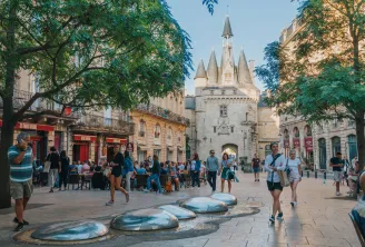Das Cailhau-Tor, ein historisches Denkmal in Bordeaux, von einem schattigen öffentlichen Platz mit Besuchern, Cafés und Bodenskulpturen aus gesehen