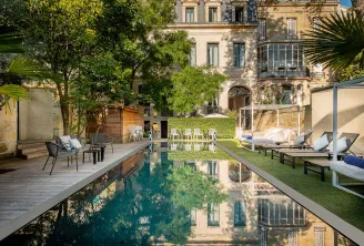 Ein rechteckiges Spiegelbecken umgeben von üppigen Grünanlagen, Liegestühlen und Entspannungsbereichen mit Blick auf die elegante klassische Fassade des Palais Gallien Hôtel & Spa in Bordeaux