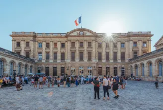 Fassade des Palais Rohan in Bordeaux mit seinem gepflasterten Hof, klassischer Steinarchitektur, französischer Flagge auf der Spitze und Besuchern im Hof an einem sonnigen Tag.