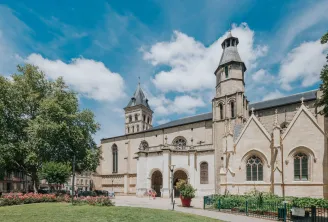 Die Basilika Saint-Seurin in Bordeaux, ein Gotteshaus aus weißem Stein mit zwei Türmen, umgeben von Grünanlagen und einem gestalteten Platz unter blauem Himmel.