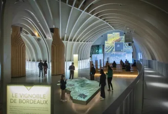Innenraum der Cité du Vin in Bordeaux mit moderner Architektur, glänzend weißem Gewölbedach, Besuchern, die ein beleuchtetes Modell des Bordeaux-Weinbergs betrachten, und Ausstellungsbildschirmen an den Wänden.