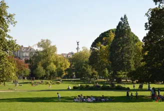 Blick auf den Öffentlichen Garten von Bordeaux mit grünen Rasenflächen, majestätischen Bäumen, entspannten Besuchern, Denkmal mit Statue im Hintergrund und historischen Gebäuden unter klarem Himmel.
