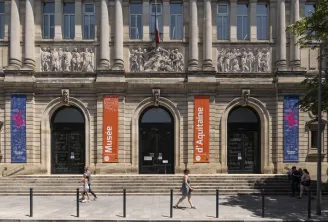 Fassade des Musée d'Aquitaine in Bordeaux mit imposanten Säulen, Reliefplatten, orange und blauen Bannern, drei Rundbogenportalen und Besuchern auf dem Platz.