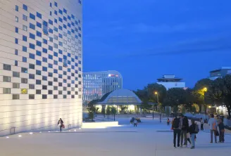 Nachtansicht der Méca in Bordeaux mit ihrer beleuchteten weißen Schachbrettfassade, gepflastertem öffentlichen Platz, Besuchern und modernen Gebäuden bei Dämmerung.