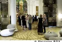 Interieur der Wine Bar des CIVB in Bordeaux mit Besuchern, umgeben von Metallausstellungspanelen, einem farbigen Buntglasfenster im Hintergrund und weißen geschwungenen Sitzmöbeln im Vordergrund.