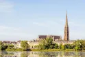 Panoramablick auf die Basilika Saint-Michel in Bordeaux mit ihrem gotischen Spitzturm, der die Stadtlandschaft dominiert, umgeben von Haussmann-Gebäuden und gespiegelt in den Gewässern der Garonne.