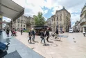 Belebter Platz in Bordeaux mit Fußgängern, Radfahrern und Geschäften, umgeben von Haussmann-Gebäuden unter bewölktem Himmel.