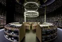Moderner Weinkeller des Latitude 20 Wine Bar in Bordeaux mit gekrümmten Regalen voller Weinflaschen, zentralem Holztresen und kreisförmiger weißer Deckenbeleuchtung.