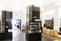 Moderner Innenraum der Max Bordeaux Wine Gallery & Cellar mit beleuchteten Weinvitrinen, stapelbaren Holzkisten und Verkostungsausrüstung in einem hellen, minimalistischen Raum.